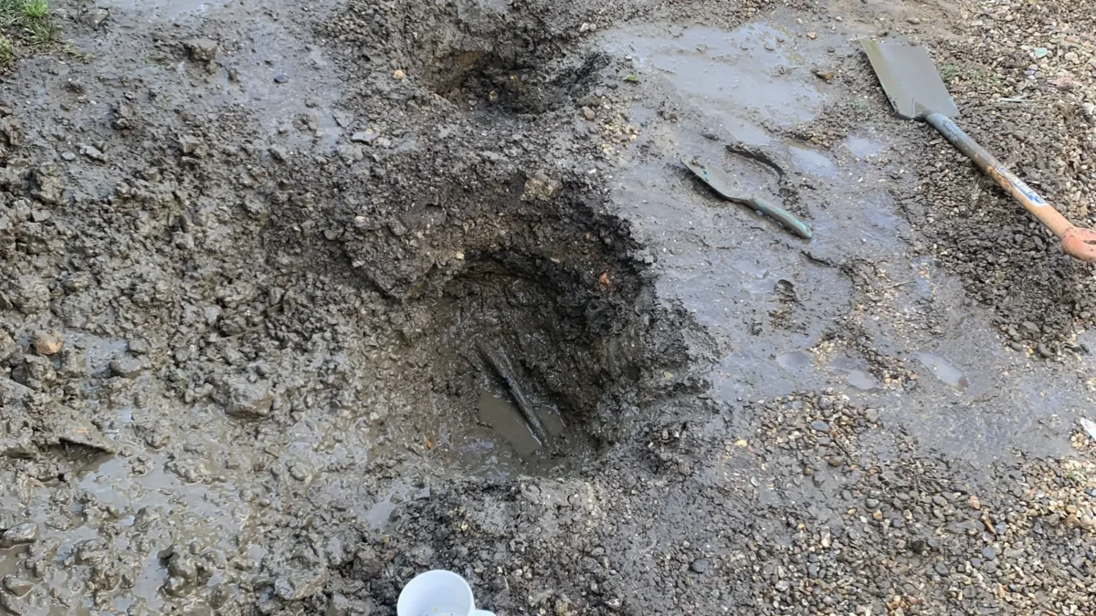 Burst alcathene water main repair excavation in North Tuddenham, Dereham, showing muddy hole with tools and mug