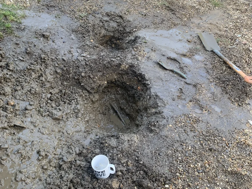 Burst alcathene water main repair excavation in North Tuddenham, Dereham, showing muddy hole with tools and mug
