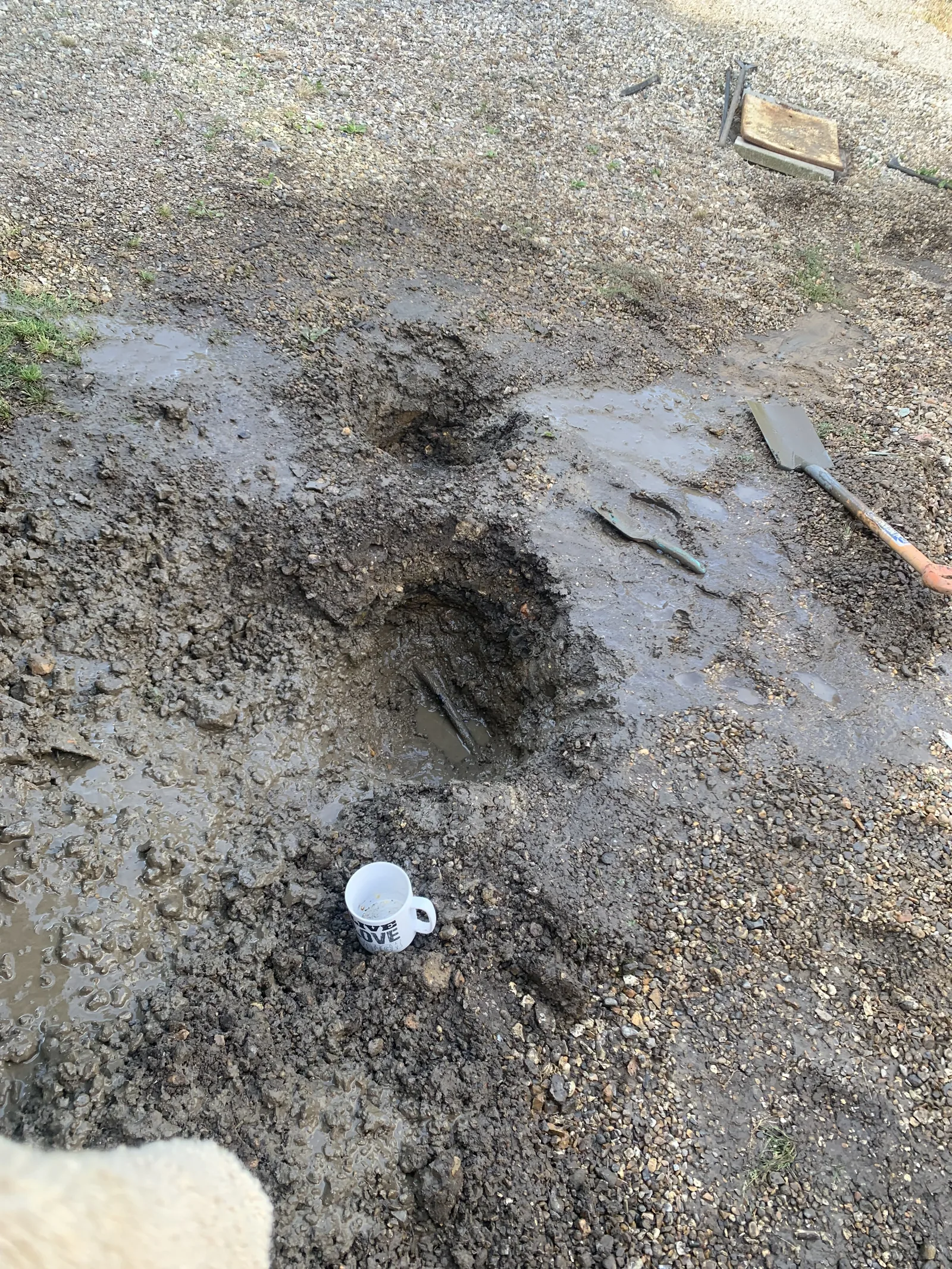 Burst alcathene water main repair excavation in North Tuddenham, Dereham, showing muddy hole with tools and mug