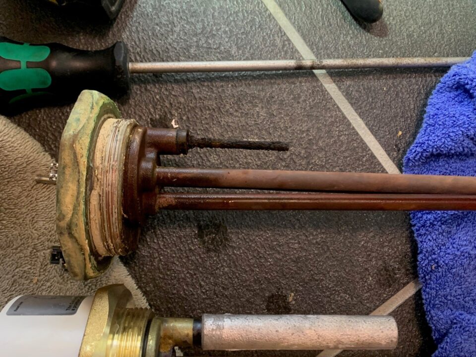 Several immersion heater elements, including one badly corroded unit, lie beside plumbing tools on a tiled floor.