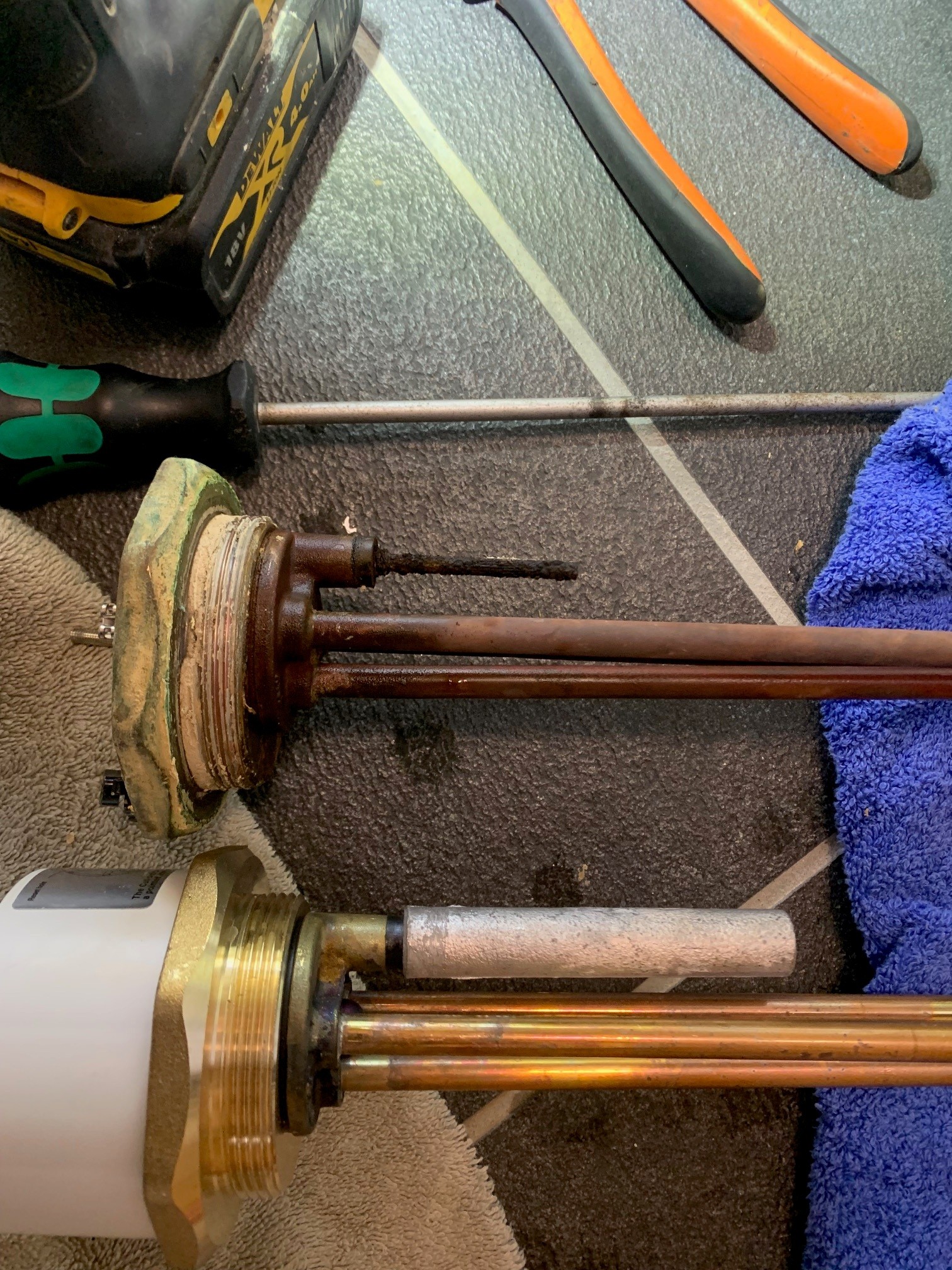 Several immersion heater elements, including one badly corroded unit, lie beside plumbing tools on a tiled floor.